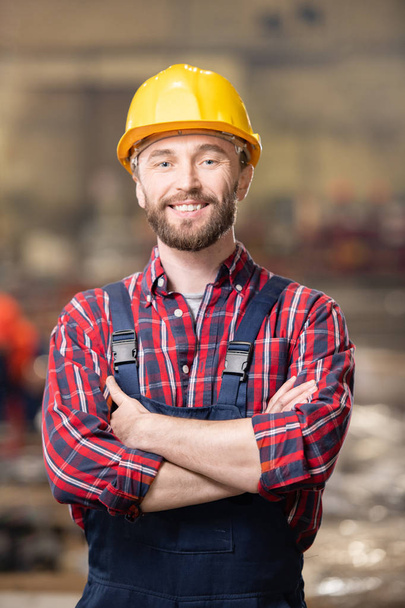 Счастливый молодой улыбающийся инженер или машиностроитель в каске и комбинезоне скрещивая руки на груди
 - Фото, изображение