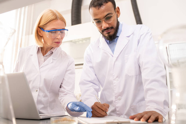 Серьезные вдумчивые многонациональные медицинские коллеги в лабораторных халатах, стоящие за столом с ноутбуком и делающие заметки в буфете во время наблюдения в лаборатории
 - Фото, изображение