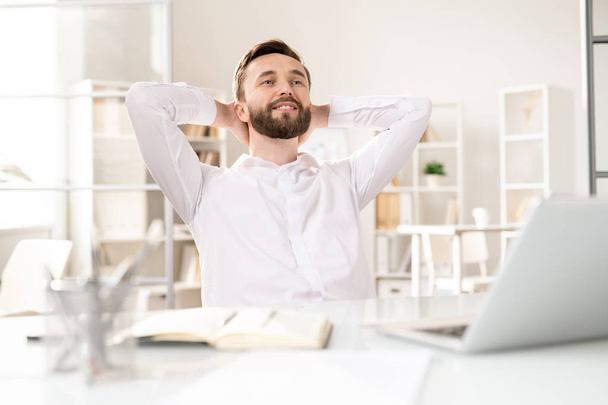 Gelukkige jonge rustgevende ondernemer zittend op werkplek met zijn handen achter hoofd en dagdromen bij pauze of gewoon krijgen geïnspireerd - Foto, afbeelding