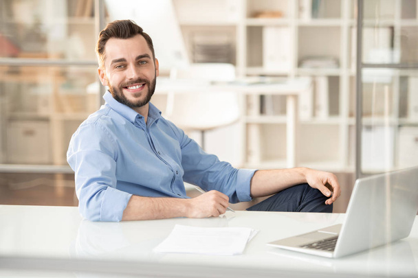 Jonge vrolijke manager met tand glimlach zitten door bureau met laptop aan de voorkant en werken over planning - Foto, afbeelding