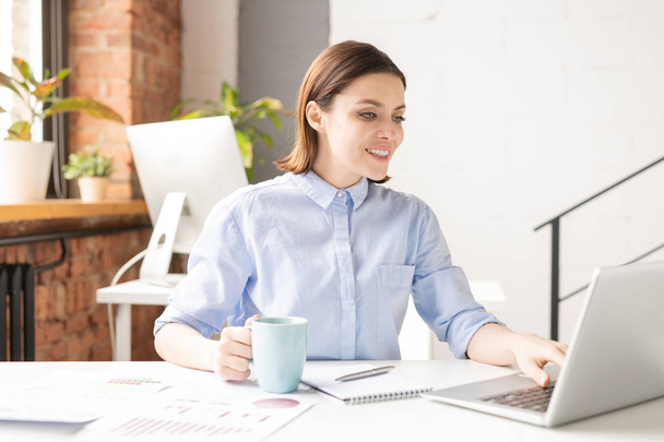 Счастливая молодая успешная женщина-брокер в рубашке смотрит на дисплей ноутбука во время серфинга в сети во время кофе-брейка в офисе
 - Фото, изображение