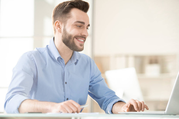 Empleado barbudo feliz en camisa azul sentado delante de la computadora portátil mientras navega en la red para obtener alguna información en particular
 - Foto, imagen