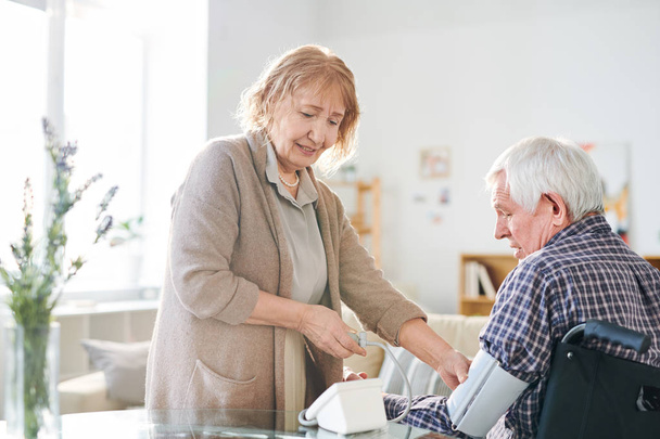 Femme âgée en tenue décontractée utilisant un tonomètre pour mesurer la pression artérielle de son mari handicapé à la maison
 - Photo, image