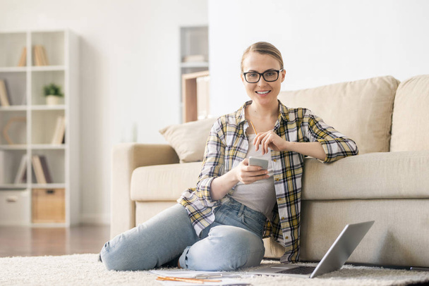 Onnellinen itsevarma nuori nainen lasit istuu matolla olohuoneessa ja katselee kameraa, kun käytät gadgetia ja ajattelet startup-ideoita
 - Valokuva, kuva