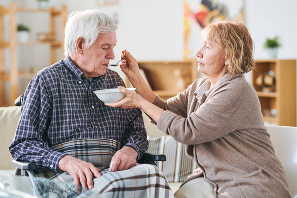 Aged casual woman holding bowl with soup and spoon by her disable husband mouth while helping him to eat dinner - Photo, Image