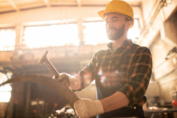 Young worker of contemporary machinebuilding plant holding metallic workpiece and hammering it - Φωτογραφία, εικόνα