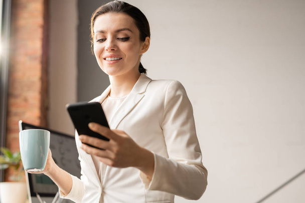 Succesvolle vrouwelijke Office Manager in wit pak lezen bericht of melding in smartphone bij koffiepauze in Office - Foto, afbeelding