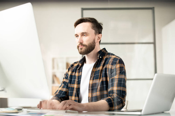 Ciddi genç webtasarımcısı bilgisayar ekranının önünde otururken online bilgi üzerinden bakarak konsantre - Fotoğraf, Görsel