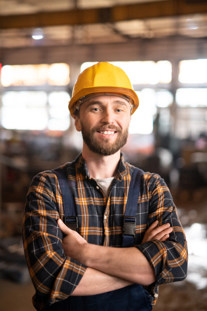 Young cheerful successful engineer or foreman in workwear and protective helmet standing in workshop in front of camera - Foto, Imagem