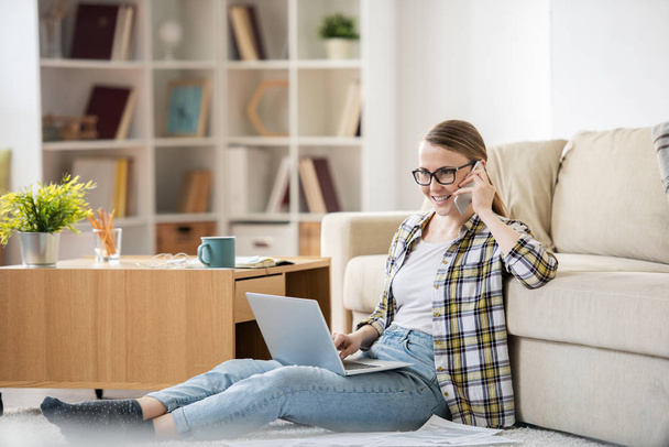 Pozytywne pewni młoda kobieta zdalnego kierownika w casual Odzież siedzi z laptopem na podłodze w salonie i rozmawiać na telefon w domu - Zdjęcie, obraz