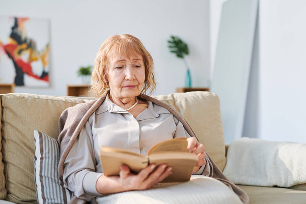 Mujer mayor en ropa casual sentada en cómodo sofá por cojín y leyendo libro de cuentos o novela en la sala de estar
 - Foto, Imagen