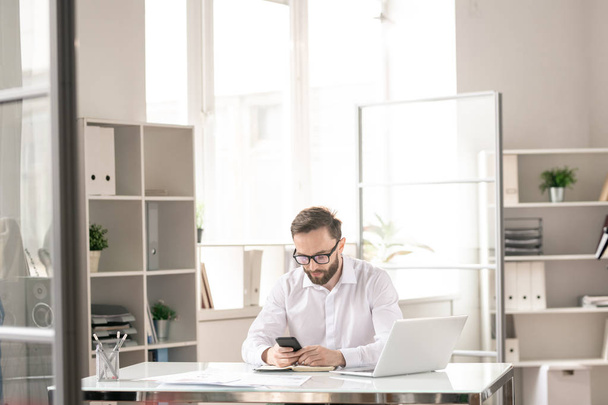 Jeune homme d'affaires contemporain assis par bureau dans le bureau et textos ou défilement dans le smartphone au travail
 - Photo, image