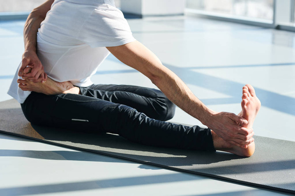 Young man in activewear sitting on mat while holding his feet by hands during stretching yoga exercise - Valokuva, kuva