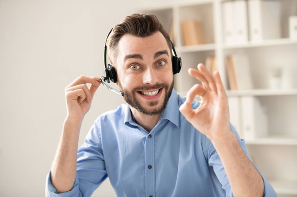 Happy young operator with toothy smile showing okay gesture while looking at you and comunicating on headphone - Foto, Imagen