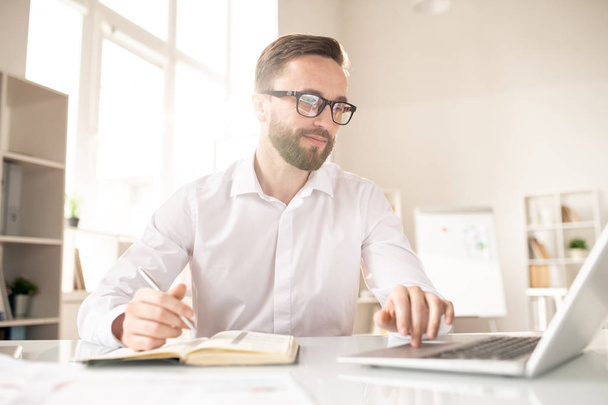 Молодой офисный менеджер или аналитик концентрируется на работе с онлайн данными, сидя за столом перед ноутбуком
 - Фото, изображение