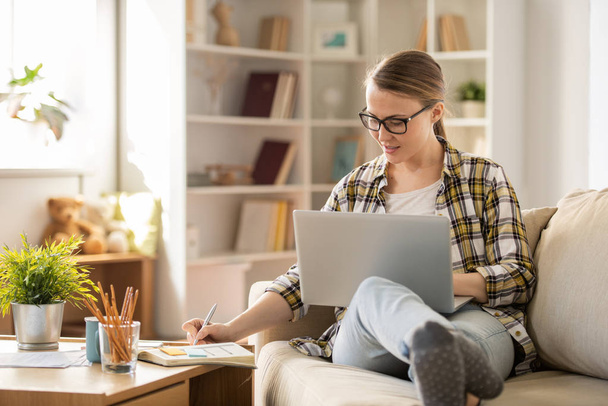 Menina atraente positivo em óculos sentados no sofá e fazendo anotações no diário enquanto trabalhava em casa
 - Foto, Imagem