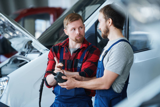 Один из молодых специалистов по ремонту автомобилей указывает на электронное устройство в руках своего коллеги во время рабочего разговора
 - Фото, изображение