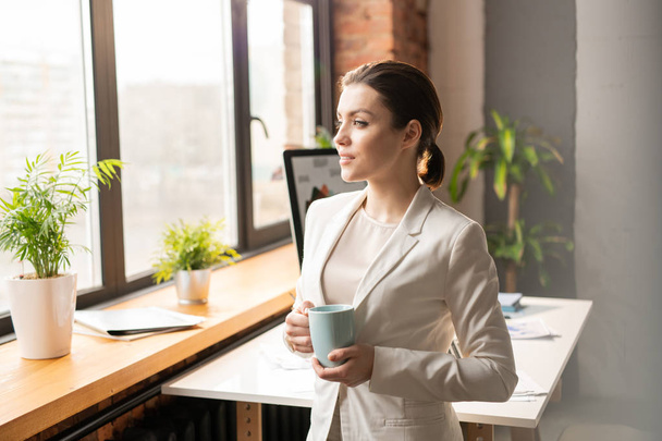 Jovem empresária encantadora em formalwear branco tomando café ou chá na pausa enquanto estiver no escritório
 - Foto, Imagem
