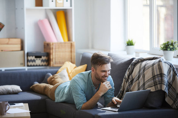 Portrait complet de beau jeune homme utilisant un ordinateur portable couché sur le canapé à la maison et souriant, espace de copie
 - Photo, image