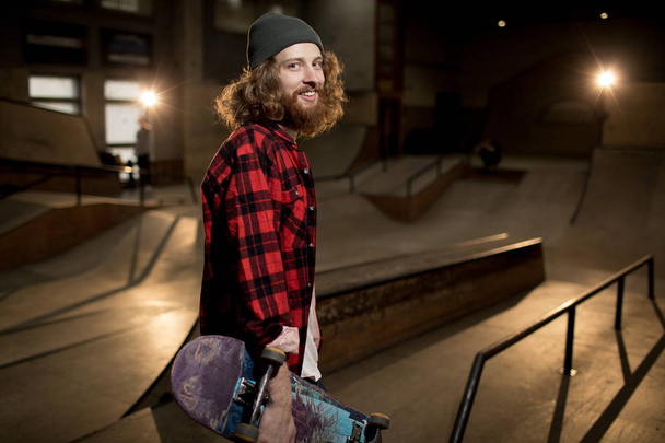 Waist up portrait of long haired man holding skateboard looking at camera standing in extreme sports park, shot with flash - Foto, Imagen