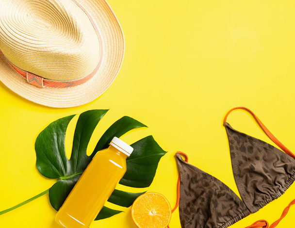 Summer beach accessories hat swimsuit bottle of orange juice bright yellow background big green leaves tropical plant. - Photo, Image