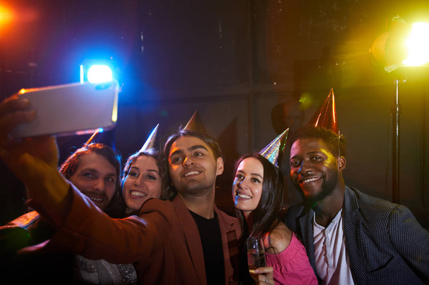 Przyjęcie urodzinowe w klubie nocnym: Grupa pozytywnych młodych wieloetnicznych przyjaciół w imprezowych kapeluszach stwarzających do selfie w ciemnym pomieszczeniu z lampami Disco - Zdjęcie, obraz