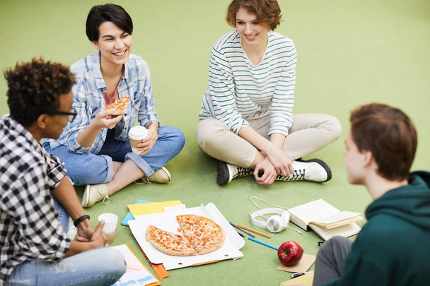Група позитивних молодих багатоетнічних студентів у повсякденному вбранні, що сидять на підлозі та їдять піцу, спілкуючись разом та сміючись
 - Фото, зображення