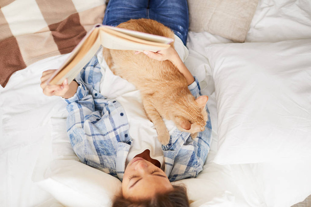 Top view portrait of young woman reading book laying in bed with ginger cat, copy space - Foto, Imagen