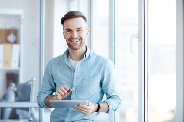Waist up portrait of handsome young man holding digital tablet and smiling happily at camera standing by window, copy space - Photo, Image