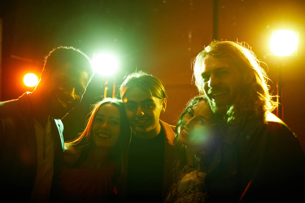 Веселые красивые молодые друзья стоят вместе в ярких огнях диско и смотрят в камеру, веселясь в ночном клубе
 - Фото, изображение