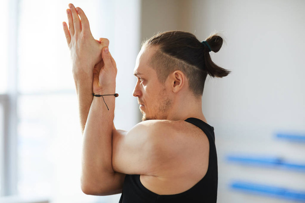 Concentrated handsome young man with ponytail performing yoga pose including eagle arms and looking straight - Fotoğraf, Görsel