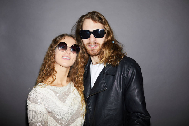 Cool fashionable young couple in sunglasses standing against gray wall and looking at camera - Φωτογραφία, εικόνα