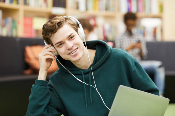 Allegro bel giovane studente maschio con cappuccio verde seduto nella zona della biblioteca e ascoltare musica in cuffie cablate mentre guarda la fotocamera
 - Foto, immagini