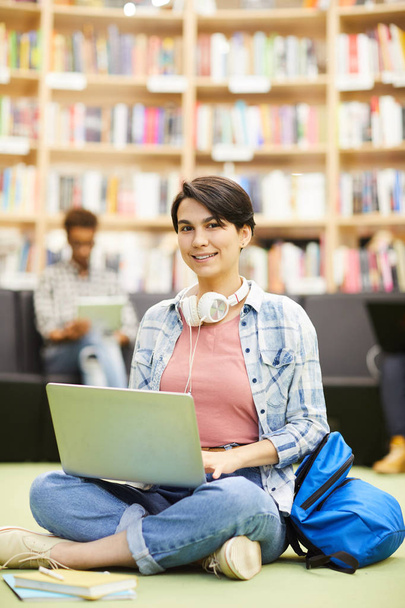 positive Hipster-Studentin in lässigem Outfit sitzt mit überkreuzten Beinen auf dem Boden in der Bibliothek und nutzt Online-Ressourcen auf dem Laptop, während sie Hausaufgaben in der Bibliothek macht. - Foto, Bild
