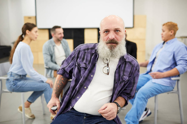Retrato del hombre mayor barbudo mirando la cámara mientras está sentado en la silla frente al grupo de terapia, espacio para copiar
 - Foto, Imagen