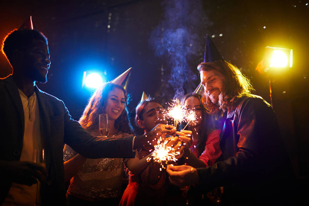 Group of happy young friends in party caps standing in circle and lighting sparklers together while preparing for surprise party - Photo, Image