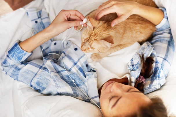 Portrait de jeune femme contemporaine couchée au lit avec un chat roux et écoutant de la musique, copiant de l'espace
 - Photo, image