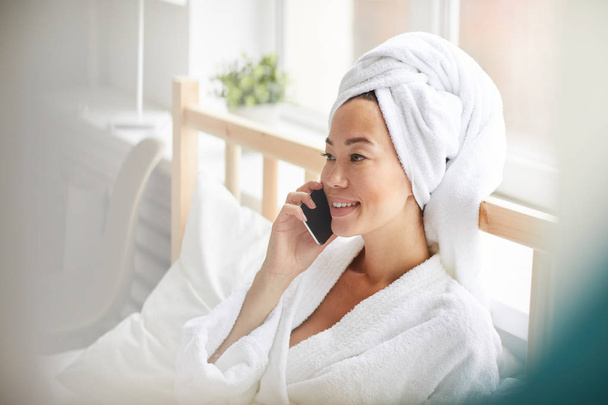 Widok z boku uśmiechnięta Azjatycka kobieta ubrana w szatę do kąpieli mówiąca przez telefon podczas relaksu w łóżku w domu, Kopiuj przestrzeń - Zdjęcie, obraz