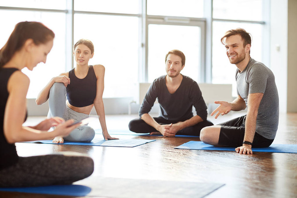 Grupo de jovens estudantes curiosos positivos sentados no chão em sala cheia de luz natural e conversando com professor de ioga no seminário
 - Foto, Imagem