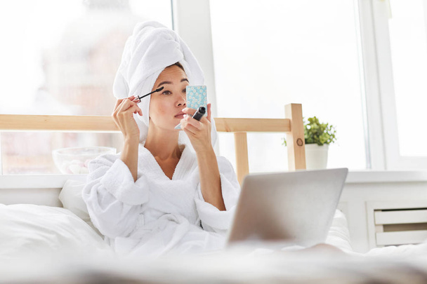 Portrait of  gorgeous Asian woman wearing bath robe doing makeup in bed  while  relaxing at home, copy space - Photo, Image