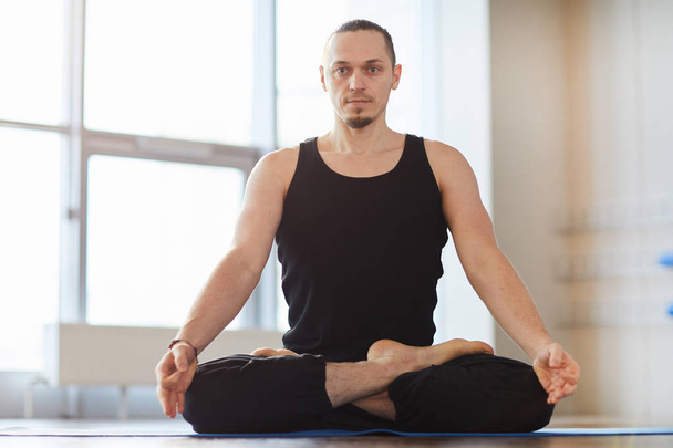 Serious muscled young man with beard sitting in lotus position and meditating alone in empty room, he looking at camera - Фото, изображение