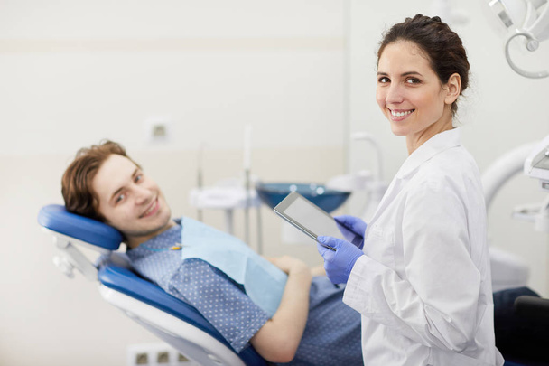 Taille up portret van vrouwelijke tandarts glimlachend gelukkig bij de camera tijdens het raadplegen van de patiënt op de achtergrond, kopieer ruimte - Foto, afbeelding