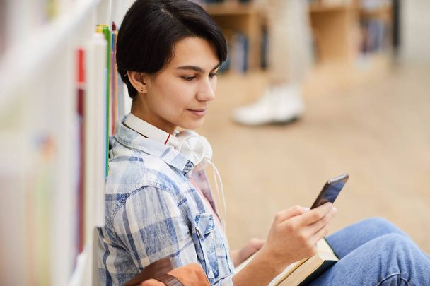 İçerik kısa siyah saçlı güzel üniversite öğrencisi kız boynunda kulaklık takan kütüphanede katta oturan ve telefonda mesaj aylaklama - Fotoğraf, Görsel