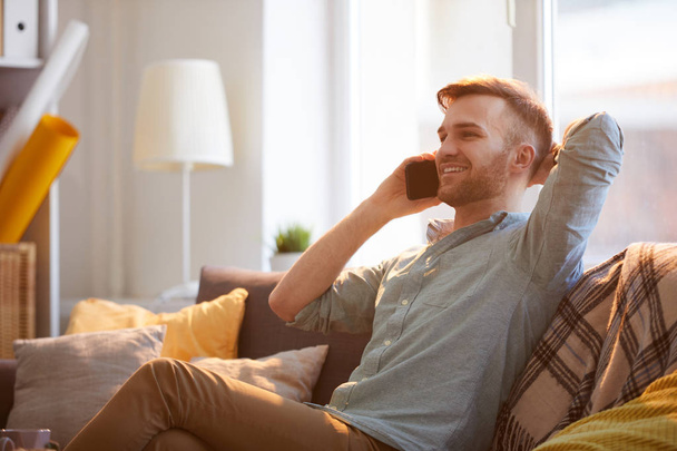 Portrait of handsome young man speaking by phone sitting on couch in sunlight, copy space - Photo, Image