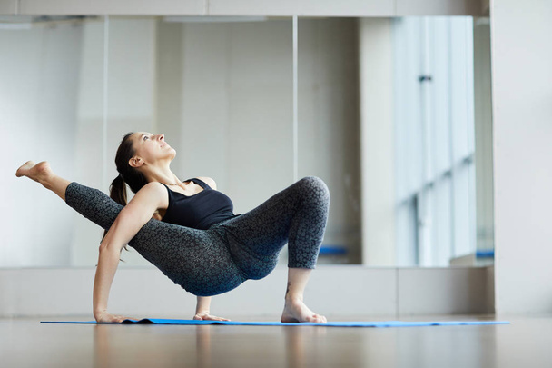 Confident attractive professional yogis in leggings and wifebeater standing on one foot and raising leg aside through arm while doing modified yoga crab pose in studio. - Foto, imagen