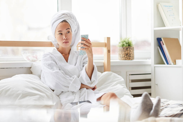 Портрет чудової азіатській жінці носіння ванни халат за допомогою смартфона, відпочиваючи в ліжку вдома, копіювати простір - Фото, зображення