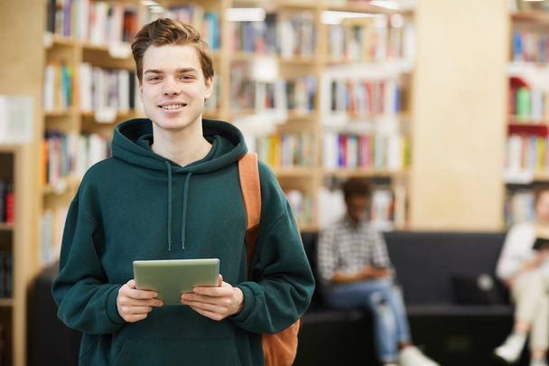 Alegre estudiante de secundaria guapo seguro de sí mismo con bolsa de pie en la biblioteca moderna y el uso de la tableta digital mientras mira a la cámara
 - Foto, imagen