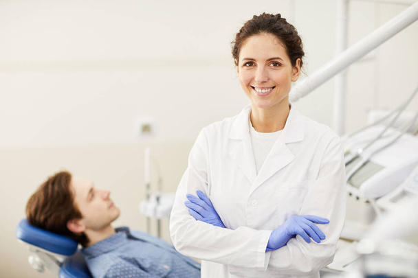 Taille up portret van vrouwelijke tandarts glimlachend op de camera tijdens het raadplegen van de patiënt op de achtergrond, kopieer ruimte - Foto, afbeelding