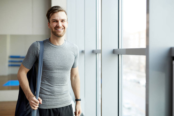 Cheerful handsome young man with stubble wearing gray tshirt standing in modern yoga studio and holding mat bag on shoulder while looking at camera - 写真・画像