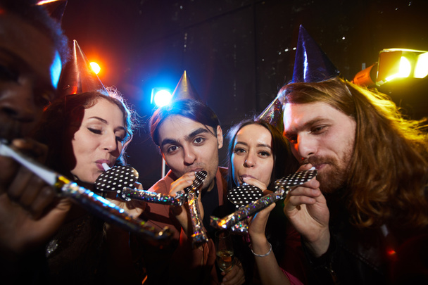 Друзья устраивают вечеринку-сюрприз: группа веселых молодых людей в партийных кепках, стоящих кругами и дующих в рога вечеринок в темной комнате
 - Фото, изображение